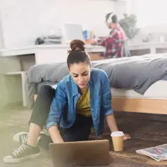 Młoda kobieta pracuje na laptopie na podłodze w pokoju. Obok niej kubek kawy i gazeta. W tle mężczyzna w słuchawkach. Zastanawia się, ile osób może mieszkać w wynajmowanym mieszkaniu.