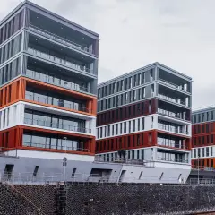 Nowoczesne budynki o geometrycznych kształtach, z elementami w kolorze pomarańczowym i bordowym, tworzą futurystyczny krajobraz miasta nad wodą.