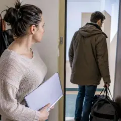 Kobieta z teczką patrzy na byłego męża wychodzącego z torbą. Sytuacja sugeruje eksmisję byłego małżonka z własnego mieszkania.