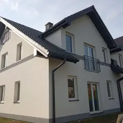 Nowy dom z jasną elewacją i ciemnym dachem. Wokół zielona rząska trawy, a na ścianach liczne okna i balkony.