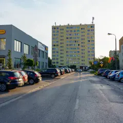 Uliczka z zaparkowanymi samochodami obok sklepu Biedronka, gdzie logo przypomina uśmiechniętego ursusa niedźwiadka. W tle wysoki, żółty blok mieszkalny.