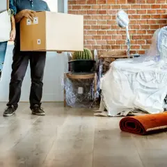 Para przeprowadzająca się, niosąca karton i kosz. Meble owinięte folią, gotowe do transportu. To ich nowy okres ochronny dla lokatorów.