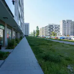 Nowoczesne osiedle w Ursusie Centralnym. Chodnik prowadzi do sklepu Żabka, a dalej widać parking z samochodami i budynki mieszkalne.