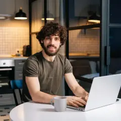Mężczyzna z kręconymi włosami pracuje na laptopie w kuchni. Analizuje koszty, porównując czynsz a wynajem mieszkania.