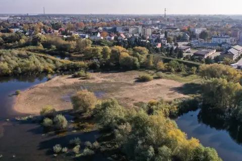 Malowniczy krajobraz z rzeką i terenami podmokłymi, w tle rozciąga się panorama jednego z miast obok Warszawy.