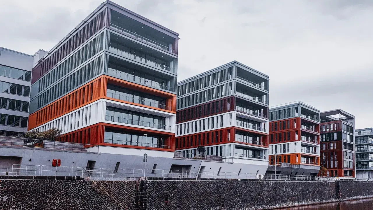 Nowoczesne budynki o geometrycznych kształtach, z elementami w kolorze pomarańczowym i bordowym, tworzą futurystyczny krajobraz miasta nad wodą.