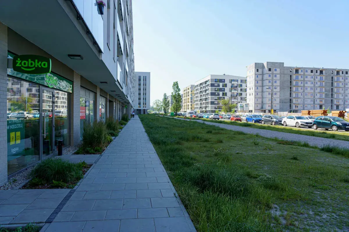 Nowoczesne osiedle w Ursusie Centralnym. Chodnik prowadzi do sklepu Żabka, a dalej widać parking z samochodami i budynki mieszkalne.