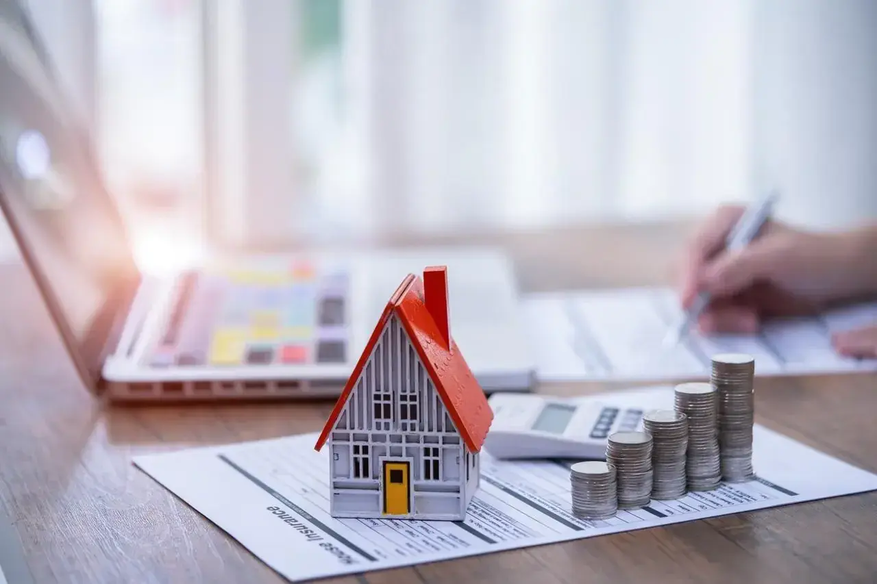 Model domu, stos monet i kalkulator na dokumencie "House Insurance". To symbol ubezpieczenia pomostowego, które chroni Twój dom.